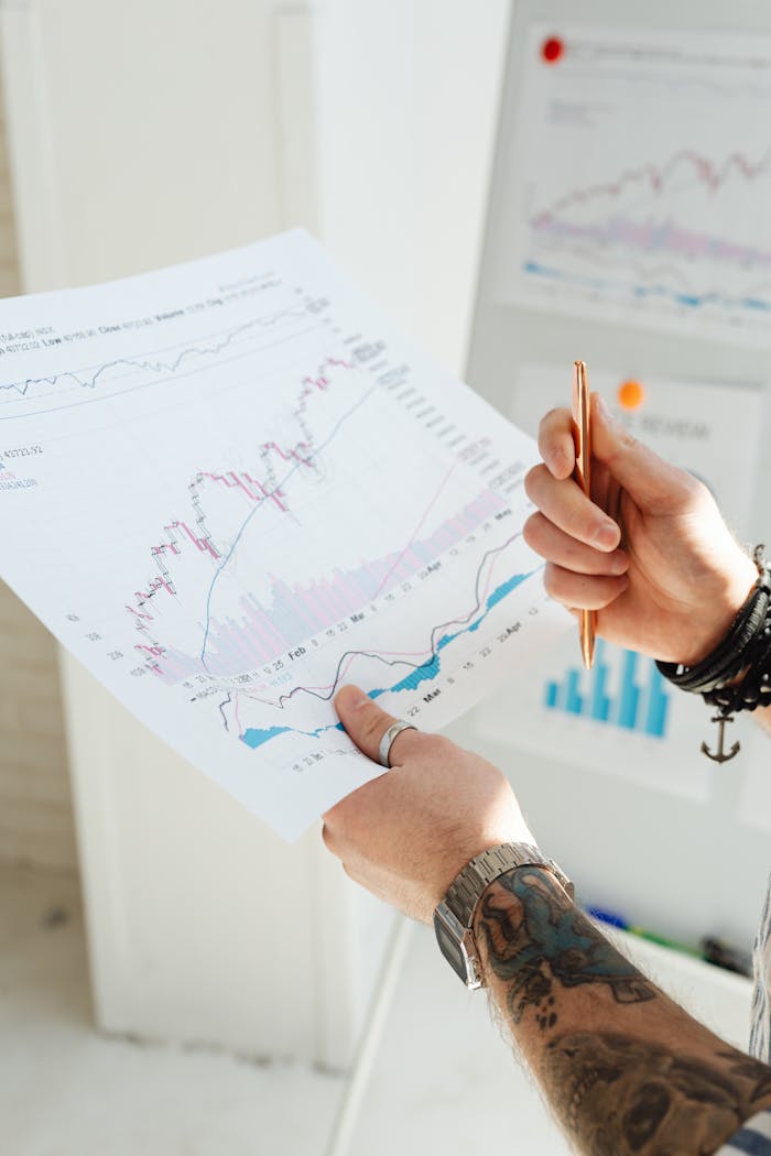 Close-up of tattooed hands holding financial charts and pen, illustrating market analysis.